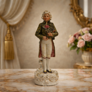 Statuina in porcellana tedesca policroma, raffigurante gentiluomo in abiti settecenteschi con parrucca incipriata e libro tra le mani, modellata a tutto tondo. Decorazione sobria ma accurata, con tocchi di colore e leggere dorature sugli abiti e sulla base sagomata a volute. Produzione Germania, fine XIX – inizi XX secolo. Normali segni del tempo, complessivamente in buono stato di conservazione.