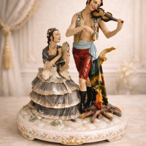 Gruppo in porcellana di Capodimonte raffigurante coppia di gitani musicanti Italia, XX secolo Raffinato gruppo scultoreo in porcellana di Capodimonte, finemente modellato e dipinto a mano, raffigurante una suggestiva scena con coppia di gitani musicanti. Il giovane violinista, in abbigliamento tradizionale con gilet decorato, fascia in vita e pantaloni al ginocchio, è colto nell’atto di suonare con espressione intensa e partecipe. Accanto a lui, la figura femminile seduta accompagna la melodia con tamburello, indossando un ampio abito a balze impreziosito da bordure policrome e scialle con frange. La composizione è arricchita da dettagli naturalistici quali il ceppo d’albero e il piccolo fuoco acceso, elementi che conferiscono dinamismo e atmosfera alla scena. Notevole la qualità della modellazione, evidente nella resa dei volti, delle mani e dei panneggi, così come nella vivace e armoniosa policromia con leggere dorature. Base sagomata decorata a rilievo. Oggetto di forte impatto decorativo, espressione della tradizione artigianale capodimontina, ideale per collezionisti e amanti della porcellana artistica italiana.