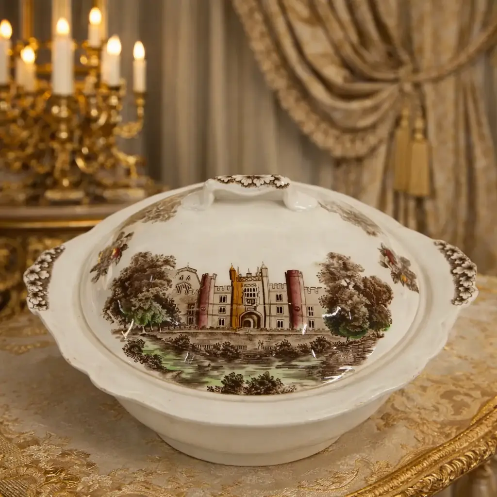 Zuppiera in ceramica inglese Johnson Bros, serie “Royal Homes of Britain – Hampton Court Palace”, manifattura Staffordshire, prima metà XX secolo. Corpo ovale con coperchio originale, decorato a trasferimento policromo raffigurante Hampton Court Palace entro paesaggio alberato, con riserve floreali sui lati e presa superiore sagomata. Marchio al verso. Buono stato di conservazione, tipico esempio di ceramica decorativa inglese di gusto storico-collezionistico.