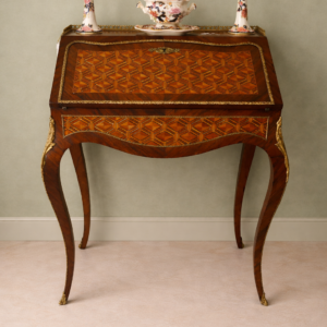 Elegante secrétaire a ribalta in stile Luigi XV, realizzato in legno massello e lastronato con pregiati intarsi geometrici a marqueterie. Il fronte presenta una raffinata ribalta sagomata decorata da motivi romboidali, incorniciata da filettature e arricchita da applicazioni in bronzo dorato. All’interno si trova un piano di scrittura rivestito in panno e una serie di piccoli cassetti impreziositi da marqueterie policrome. Il piano superiore è corredato da una galleria traforata in bronzo, mentre la base poggia su slanciate gambe mosse terminate con calzature in bronzo. Mobile di manifattura di qualità, del XIX secolo, in ottimo stato di conservazione, ideale per ambienti classici ed eleganti.