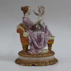 Raffinata scultura in porcellana policroma tedesca, raffigurante una giovane madre seduta su elegante seduta imbottita mentre solleva teneramente il proprio bambino, colto in un gesto affettuoso e dinamico. La scena, di gusto spiccatamente romantico, è caratterizzata da un’attenta resa anatomica, volti delicati e panneggi morbidi, impreziositi da decorazioni floreali dipinte a mano in tonalità violacee e piccoli tocchi di blu. La base sagomata e riccamente modellata presenta profili mossi con filettature dorate e finitura degradé, conferendo alla composizione un aspetto sontuoso. Sul retro del mobile compaiono ulteriori decori floreali monocromi blu, indice di una lavorazione curata anche nelle parti non immediatamente visibili. Sul fondo è presente il marchio “Dresden – Germany” con lettera K coronata, riconducibile a manifattura tedesca operante nella prima metà del XX secolo, specializzata in figure decorative di alta qualità ispirate al repertorio ottocentesco. Esemplare di notevole eleganza e forte valenza decorativa, ideale per collezionisti di porcellane europee e per ambienti classici o eclettici. Condizioni generali ottime.