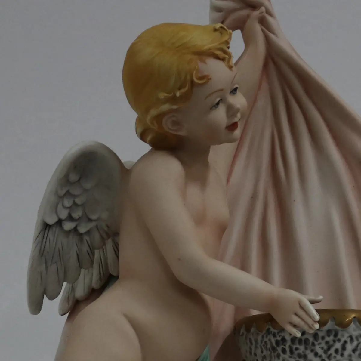 Coppia di acquasantiere da parete in porcellana policroma, raffiguranti putti alati modellati a tutto rilievo, sorreggenti vaschette con bordi dorati. Le figure, animate da movimento elegante e panneggi mossi in delicate tonalità pastello, richiamano il gusto tardo barocco–rococò reinterpretato in chiave novecentesca. Produzione italiana, manifattura Corti, XX secolo, destinate all’arredo devozionale domestico; in buono stato di conservazione.