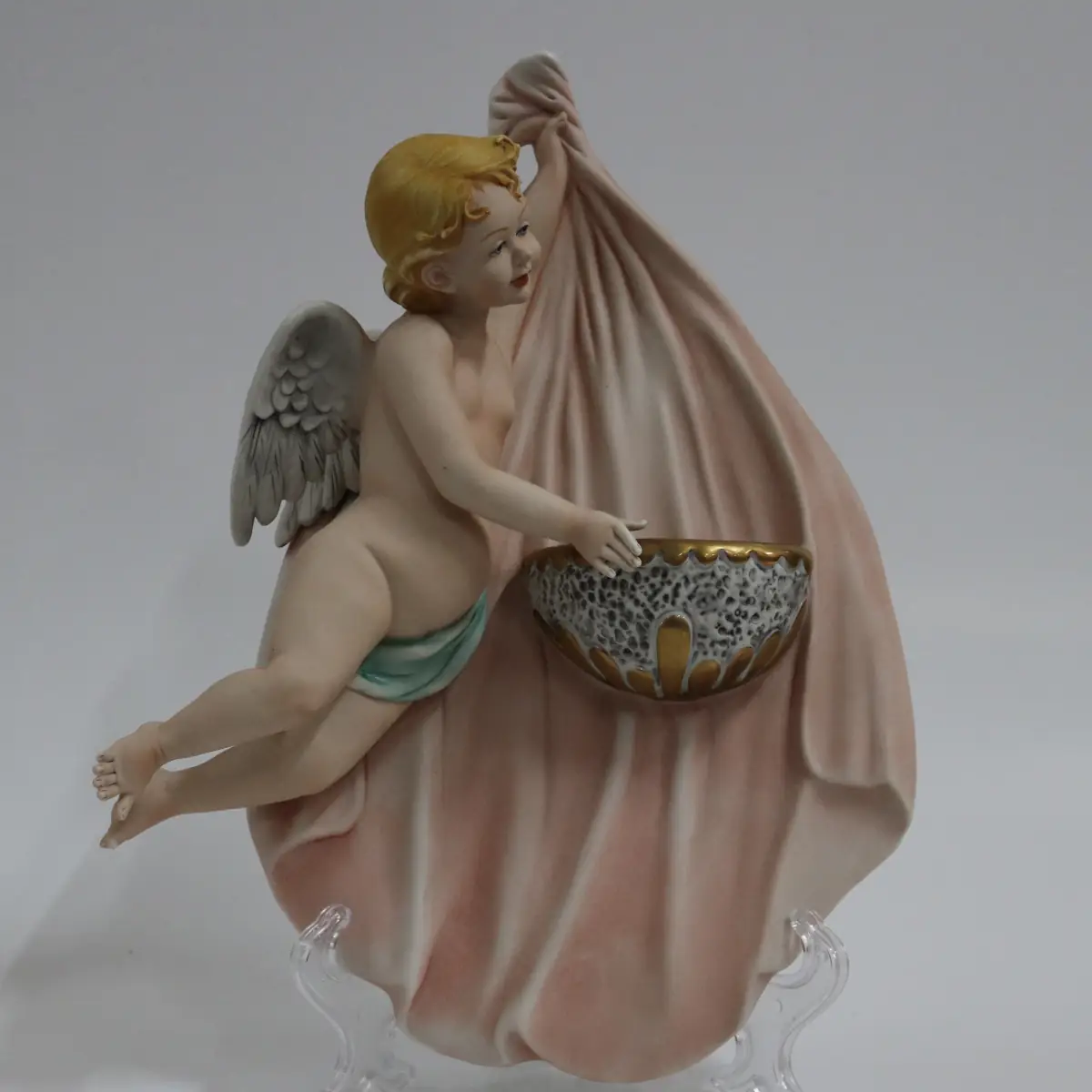 Coppia di acquasantiere da parete in porcellana policroma, raffiguranti putti alati modellati a tutto rilievo, sorreggenti vaschette con bordi dorati. Le figure, animate da movimento elegante e panneggi mossi in delicate tonalità pastello, richiamano il gusto tardo barocco–rococò reinterpretato in chiave novecentesca. Produzione italiana, manifattura Corti, XX secolo, destinate all’arredo devozionale domestico; in buono stato di conservazione.