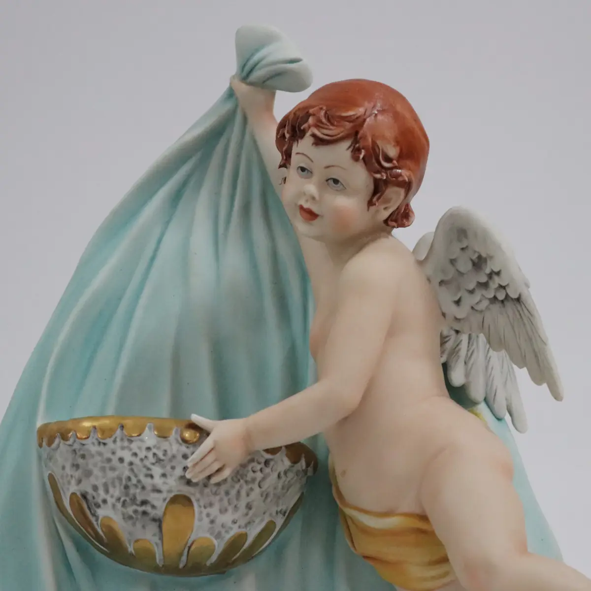Coppia di acquasantiere da parete in porcellana policroma, raffiguranti putti alati modellati a tutto rilievo, sorreggenti vaschette con bordi dorati. Le figure, animate da movimento elegante e panneggi mossi in delicate tonalità pastello, richiamano il gusto tardo barocco–rococò reinterpretato in chiave novecentesca. Produzione italiana, manifattura Corti, XX secolo, destinate all’arredo devozionale domestico; in buono stato di conservazione.