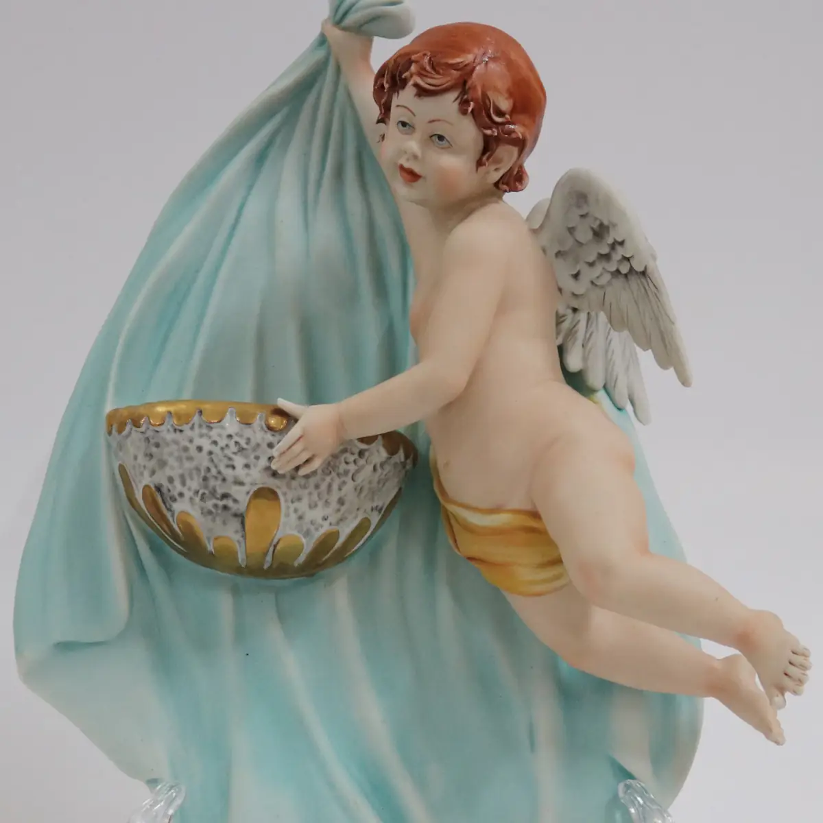 Coppia di acquasantiere da parete in porcellana policroma, raffiguranti putti alati modellati a tutto rilievo, sorreggenti vaschette con bordi dorati. Le figure, animate da movimento elegante e panneggi mossi in delicate tonalità pastello, richiamano il gusto tardo barocco–rococò reinterpretato in chiave novecentesca. Produzione italiana, manifattura Corti, XX secolo, destinate all’arredo devozionale domestico; in buono stato di conservazione.