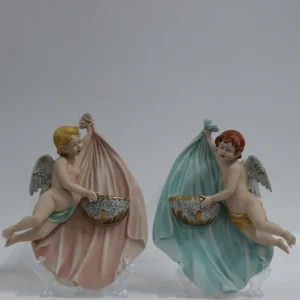 Coppia di acquasantiere da parete in porcellana policroma, raffiguranti putti alati modellati a tutto rilievo, sorreggenti vaschette con bordi dorati. Le figure, animate da movimento elegante e panneggi mossi in delicate tonalità pastello, richiamano il gusto tardo barocco–rococò reinterpretato in chiave novecentesca. Produzione italiana, manifattura Corti, XX secolo, destinate all’arredo devozionale domestico; in buono stato di conservazione.