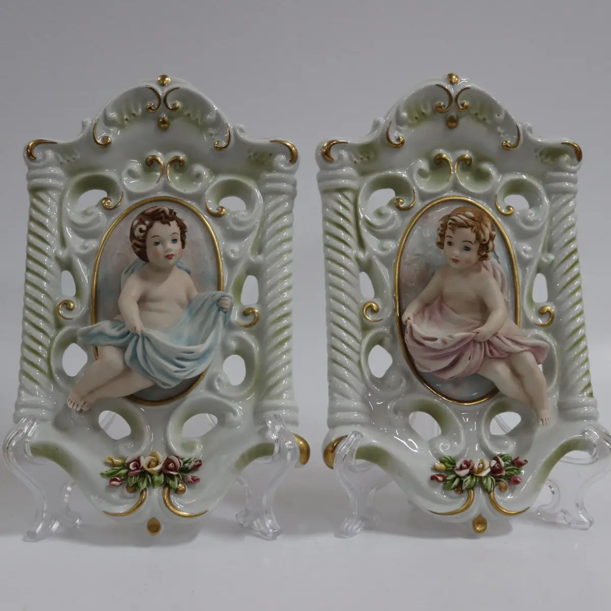 Coppia di acquasantiere da parete in porcellana policroma, in stile rococò, con struttura traforata a volute e dorature a rilievo. Al centro, putti modellati a tutto tondo con drappi azzurro e rosato entro riserva ovale. Vaschetta inferiore integrata, decorata con fiori applicati. Produzione italiana Capodimonte, XX secolo, come da marchio con N coronata e firma Corti. In buono stato di conservazione, pregevoli per arredo devozionale e collezione.