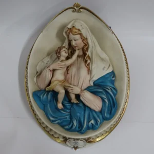 La Madonna con Bambino in porcellana di Capodimonte è un capolavoro artistico, firmato dal Maestro Corti. Quest’opera ovale da parete viene comunemente chiamata Capezzale, creata negli anni ’90 e originariamente distribuita dalla celebre manifattura artigianale King’s, ormai chiusa, è una rarità molto ricercata dai collezionisti di arte sacra. Il bassorilievo rappresenta una scena piena di dolcezza e armonia. La cura nei dettagli si riflette nella perfetta lavorazione delle mani e dei piedi del Bambino, mentre i volti sono stati delicatamente dipinti. I colori decisi e sfumati, con predominanza di azzurro, turchese e rosa. L’ovale è finemente decorato lateralmente con eleganti rifiniture in oro, che rende questa porcellana un’opera d’arte di pregio. La combinazione di dettagli raffinati e l’abilità manuale degli artigiani fanno di questa creazione una rappresentazione perfetta della porcellana di Capodimonte, che da sempre è sinonimo di alta qualità artistica e artigianale. Misure: cm. 47 x 35 x 5 Condizioni perfette