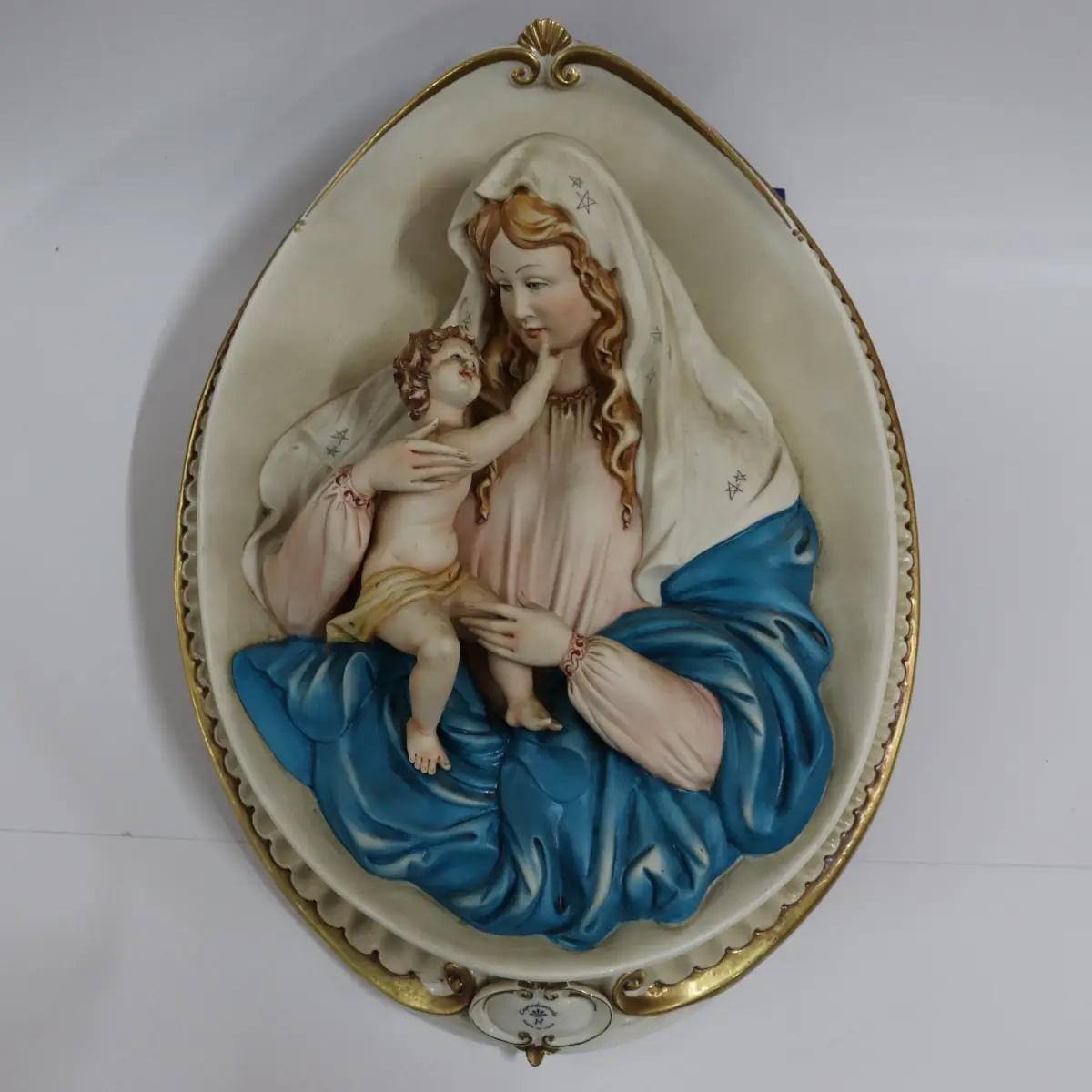 La Madonna con Bambino in porcellana di Capodimonte è un capolavoro artistico, firmato dal Maestro Corti. Quest’opera ovale da parete viene comunemente chiamata Capezzale, creata negli anni ’90 e originariamente distribuita dalla celebre manifattura artigianale King’s, ormai chiusa, è una rarità molto ricercata dai collezionisti di arte sacra. Il bassorilievo rappresenta una scena piena di dolcezza e armonia. La cura nei dettagli si riflette nella perfetta lavorazione delle mani e dei piedi del Bambino, mentre i volti sono stati delicatamente dipinti. I colori decisi e sfumati, con predominanza di azzurro, turchese e rosa. L’ovale è finemente decorato lateralmente con eleganti rifiniture in oro, che rende questa porcellana un’opera d’arte di pregio. La combinazione di dettagli raffinati e l’abilità manuale degli artigiani fanno di questa creazione una rappresentazione perfetta della porcellana di Capodimonte, che da sempre è sinonimo di alta qualità artistica e artigianale. Misure: cm. 47 x 35 x 5 Condizioni perfette