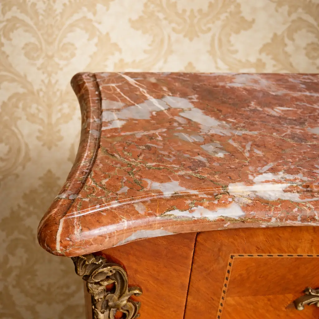 Cassettone bombato francese del XIX secolo con piano in marmo rosso Raffinato cassettone francese dell’Ottocento in stile Luigi XV, caratterizzato da linea mossa e fronte bombato. Realizzato in legni pregiati con impiallacciatura e riserve a intarsio geometrico, presenta tre ampi cassetti impreziositi da maniglie e bocchette in bronzo cesellato, in armonia con le applicazioni angolari e i sabot delle gambe.