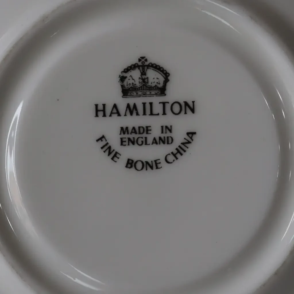 Tazza da tè con piattino in fine bone china inglese Hamilton, produzione prima metà del Novecento. Decorazione a rami e foglie stilizzate nei toni del giallo e nero, con filettature in oro e campiture in verde pallido. Forma svasata con orlo ondulato, piattino coordinato a bordo mosso. Marchio al verso: Hamilton – Made in England – Fine Bone China. In ottime condizioni, con lievi segni d’uso. Elegante oggetto da collezione o da utilizzo.