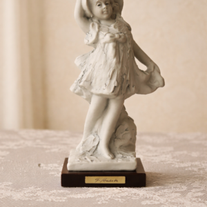 Scultura decorativa raffigurante fanciulla danzante, realizzata in bisquit con patina chiara, modellata con gusto mosso e vivace, tipico della produzione tardo Ottocento – primo Novecento. Figura stante su base naturalistica, caratterizzata da panneggio fluido e posa dinamica. Firmata “G. Bazziche” alla base; montata su basamento ligneo con targhetta. Oggetto di gusto Liberty/fin-de-siècle, destinato all’arredo borghese, in buono stato.