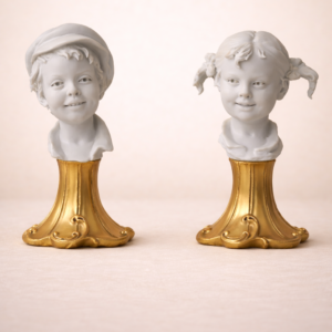 Coppia di mezzi busti raffiguranti giovani fanciulli, realizzati in porcellana di Capodimonte, finemente modellati con grande attenzione ai dettagli fisionomici ed espressivi. I volti, vivaci e sorridenti, presentano acconciature differenti che caratterizzano i soggetti, resi con naturalezza e delicatezza tipiche della migliore tradizione figurativa italiana. Entrambi i mezzi busti sono montati su basi sagomate e dorate, di gusto rocaille, che conferiscono eleganza e armonia all’insieme. Produzione italiana, XX secolo. Stato di conservazione molto buono.