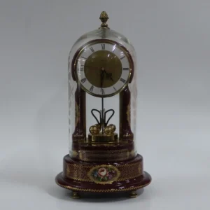 Sveglia da tavolo con movimento meccanico, racchiusa entro cupola in vetro soffiato, produzione Limoges de Sèvres, con elegante struttura in porcellana laccata color bordeaux riccamente decorata a motivi floreali policromi e dorature a mano. Base sagomata a gradini con preziose fregiature dorate e piedini in metallo. Quadrante circolare con numeri romani, fondo dorato inciso e lancette sagomate, integrato in montatura metallica. Elementi interni in metallo dorato con bilanciere decorativo. Esemplare di gusto classicheggiante, databile alla seconda metà del XX secolo, in buono stato di conservazione, completo della sua campana in vetro originale.