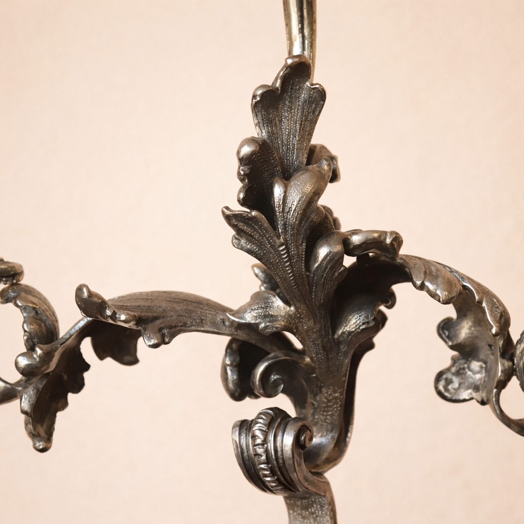 Elegante candelabro a tre luci in argento titolo 800, finemente fuso e cesellato, caratterizzato da un raffinato repertorio decorativo a volute, foglie d’acanto e motivi floreali di gusto Rococò revival. Il fusto centrale sinuoso sostiene tre bracci mossi, ciascuno concluso da portacandela a corolla, mentre la base, ampia e riccamente lavorata, garantisce equilibrio e slancio all’insieme. Databile tra la fine del XIX e i primi decenni del XX secolo, presenta una piacevole patina d’epoca ed è in buone condizioni generali. Oggetto di forte impatto decorativo, ideale per collezionisti e ambienti classici.