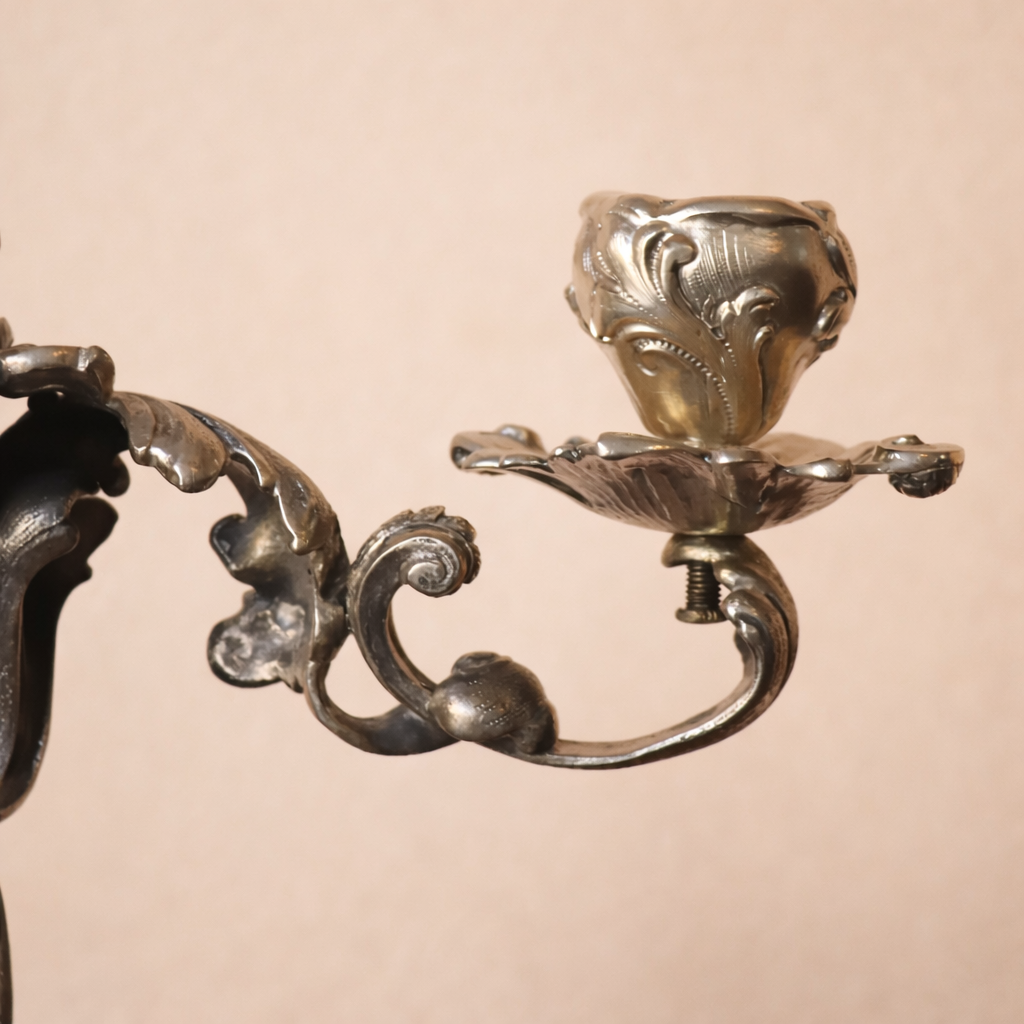 Elegante candelabro a tre luci in argento titolo 800, finemente fuso e cesellato, caratterizzato da un raffinato repertorio decorativo a volute, foglie d’acanto e motivi floreali di gusto Rococò revival. Il fusto centrale sinuoso sostiene tre bracci mossi, ciascuno concluso da portacandela a corolla, mentre la base, ampia e riccamente lavorata, garantisce equilibrio e slancio all’insieme. Databile tra la fine del XIX e i primi decenni del XX secolo, presenta una piacevole patina d’epoca ed è in buone condizioni generali. Oggetto di forte impatto decorativo, ideale per collezionisti e ambienti classici.