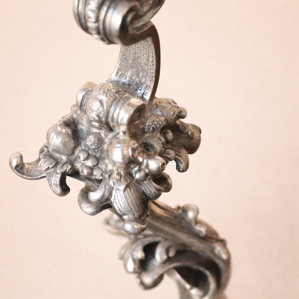 Elegante candelabro a tre luci in argento titolo 800, finemente fuso e cesellato, caratterizzato da un raffinato repertorio decorativo a volute, foglie d’acanto e motivi floreali di gusto Rococò revival. Il fusto centrale sinuoso sostiene tre bracci mossi, ciascuno concluso da portacandela a corolla, mentre la base, ampia e riccamente lavorata, garantisce equilibrio e slancio all’insieme. Databile tra la fine del XIX e i primi decenni del XX secolo, presenta una piacevole patina d’epoca ed è in buone condizioni generali. Oggetto di forte impatto decorativo, ideale per collezionisti e ambienti classici.