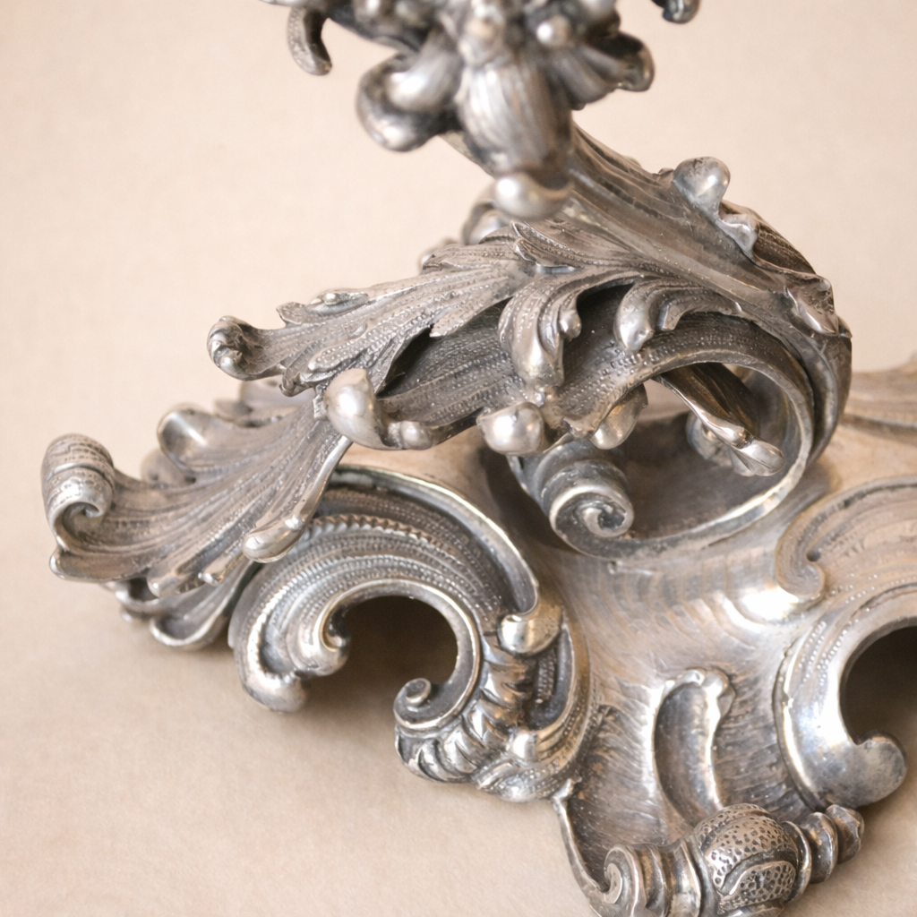Elegante candelabro a tre luci in argento titolo 800, finemente fuso e cesellato, caratterizzato da un raffinato repertorio decorativo a volute, foglie d’acanto e motivi floreali di gusto Rococò revival. Il fusto centrale sinuoso sostiene tre bracci mossi, ciascuno concluso da portacandela a corolla, mentre la base, ampia e riccamente lavorata, garantisce equilibrio e slancio all’insieme. Databile tra la fine del XIX e i primi decenni del XX secolo, presenta una piacevole patina d’epoca ed è in buone condizioni generali. Oggetto di forte impatto decorativo, ideale per collezionisti e ambienti classici.