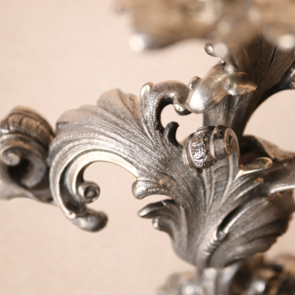 Elegante candelabro a tre luci in argento titolo 800, finemente fuso e cesellato, caratterizzato da un raffinato repertorio decorativo a volute, foglie d’acanto e motivi floreali di gusto Rococò revival. Il fusto centrale sinuoso sostiene tre bracci mossi, ciascuno concluso da portacandela a corolla, mentre la base, ampia e riccamente lavorata, garantisce equilibrio e slancio all’insieme. Databile tra la fine del XIX e i primi decenni del XX secolo, presenta una piacevole patina d’epoca ed è in buone condizioni generali. Oggetto di forte impatto decorativo, ideale per collezionisti e ambienti classici.