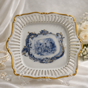 Piatto in porcellana traforata dipinta in blu cobalto, Atelier Porcelaine d’Art, XX secolo. Forma quadrilobata con tesa traforata e bordi dorati; al centro scena figurata con pescatori, ispirata alla tradizione settecentesca di Dresda e Meissen, resa in monocromia blu. Retro con iscrizioni storicizzanti e marchio dell’atelier. Buono stato di conservazione. Oggetto decorativo di gusto neoclassico, da collezione.