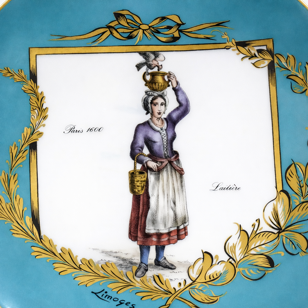 Piatto decorativo in porcellana di Limoges, con scena figurativa storica raffigurante una laitière parigina del 1600, entro cornice dorata su fondo turchese. Bordo rifinito in oro, manifattura di gusto antiquario, elegante e collezionistica.