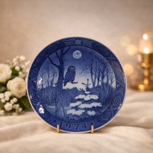 Piatto commemorativo natalizio in porcellana – Royal Copenhagen, 1974 Piatto da collezione in porcellana smaltata a decorazione monocroma blu sottocobalto, prodotto dalla manifattura Royal Copenhagen. Appartenente alla serie natalizia annuale, reca la data 1974 in rilievo sul fronte. La decorazione centrale raffigura un paesaggio boschivo invernale notturno con gufo posato su un ramo, abete innevato e luna piena, reso con eleganti sfumature di blu e atmosfera contemplativa. Bordo decorato a rilievo con motivi vegetali e stella simbolica. Al verso, marchio originale Royal Copenhagen con corona reale, tre onde e dicitura Copenhagen – Denmark, accompagnato dal titolo bilingue “Jul på Slottet / Christmas at the Manor House”.