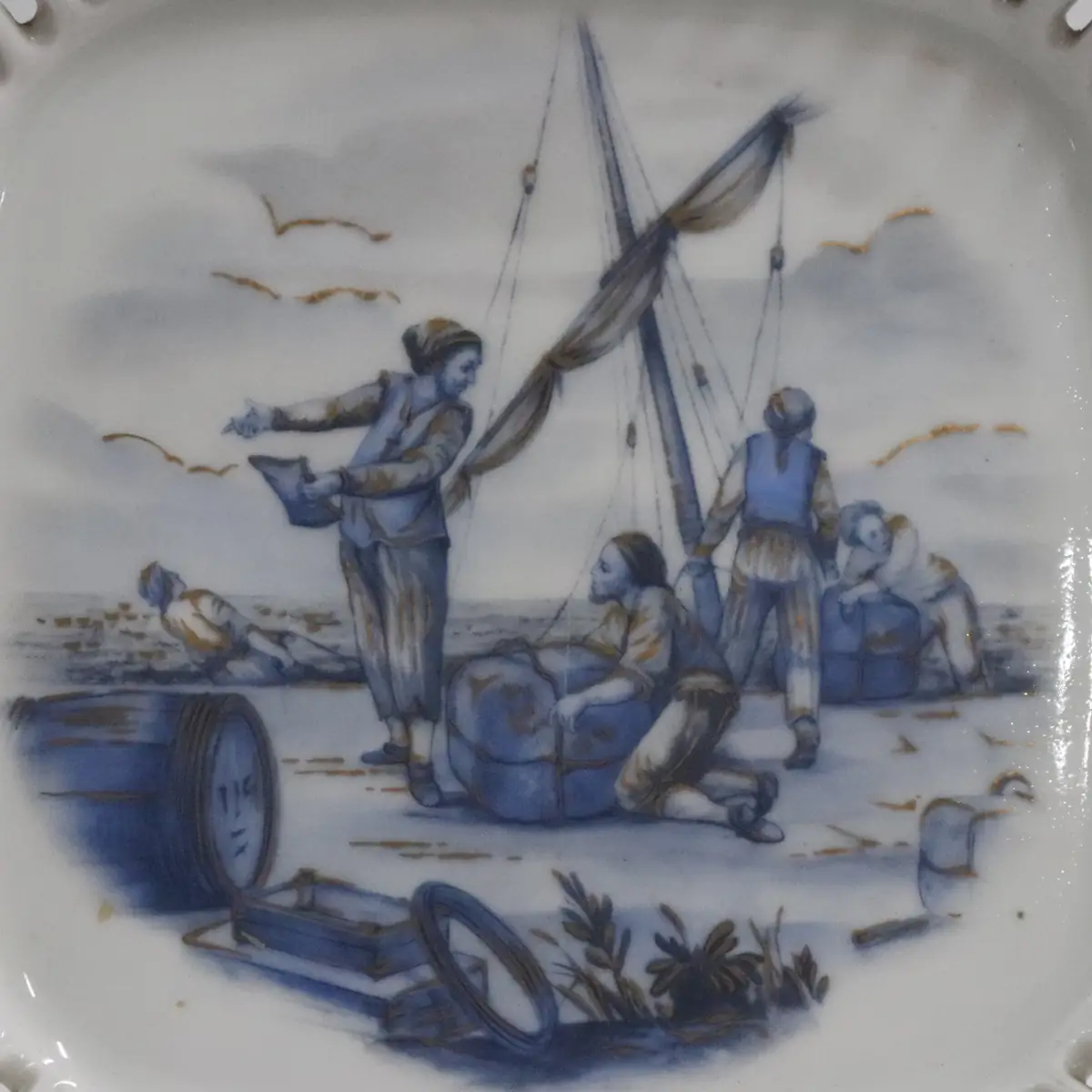 Piatto in porcellana europea del XX secolo, di gusto storico-decorativo, con scena centrale in blu cobalto raffigurante pescatori intenti alle operazioni di carico presso una barca, ispirata alle incisioni e alle manifatture francesi del Settecento. Bordo sagomato traforato con profilo dorato, esecuzione accurata e pittura sottovetrina. Il retro reca marchi e iscrizioni celebrative riferite alla Compagnia delle Indie e alla tradizione di Sèvres, indicativi di produzione commemorativa o da collezione. In buono stato di conservazione, oggetto decorativo di gusto antiquario.