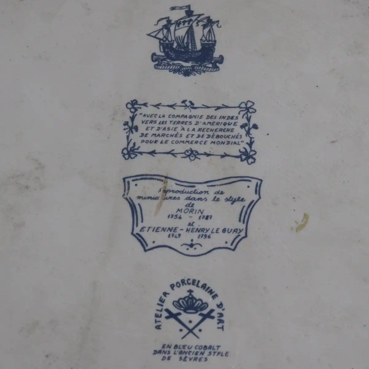 Piatto in porcellana europea del XX secolo, di gusto storico-decorativo, con scena centrale in blu cobalto raffigurante pescatori intenti alle operazioni di carico presso una barca, ispirata alle incisioni e alle manifatture francesi del Settecento. Bordo sagomato traforato con profilo dorato, esecuzione accurata e pittura sottovetrina. Il retro reca marchi e iscrizioni celebrative riferite alla Compagnia delle Indie e alla tradizione di Sèvres, indicativi di produzione commemorativa o da collezione. In buono stato di conservazione, oggetto decorativo di gusto antiquario.