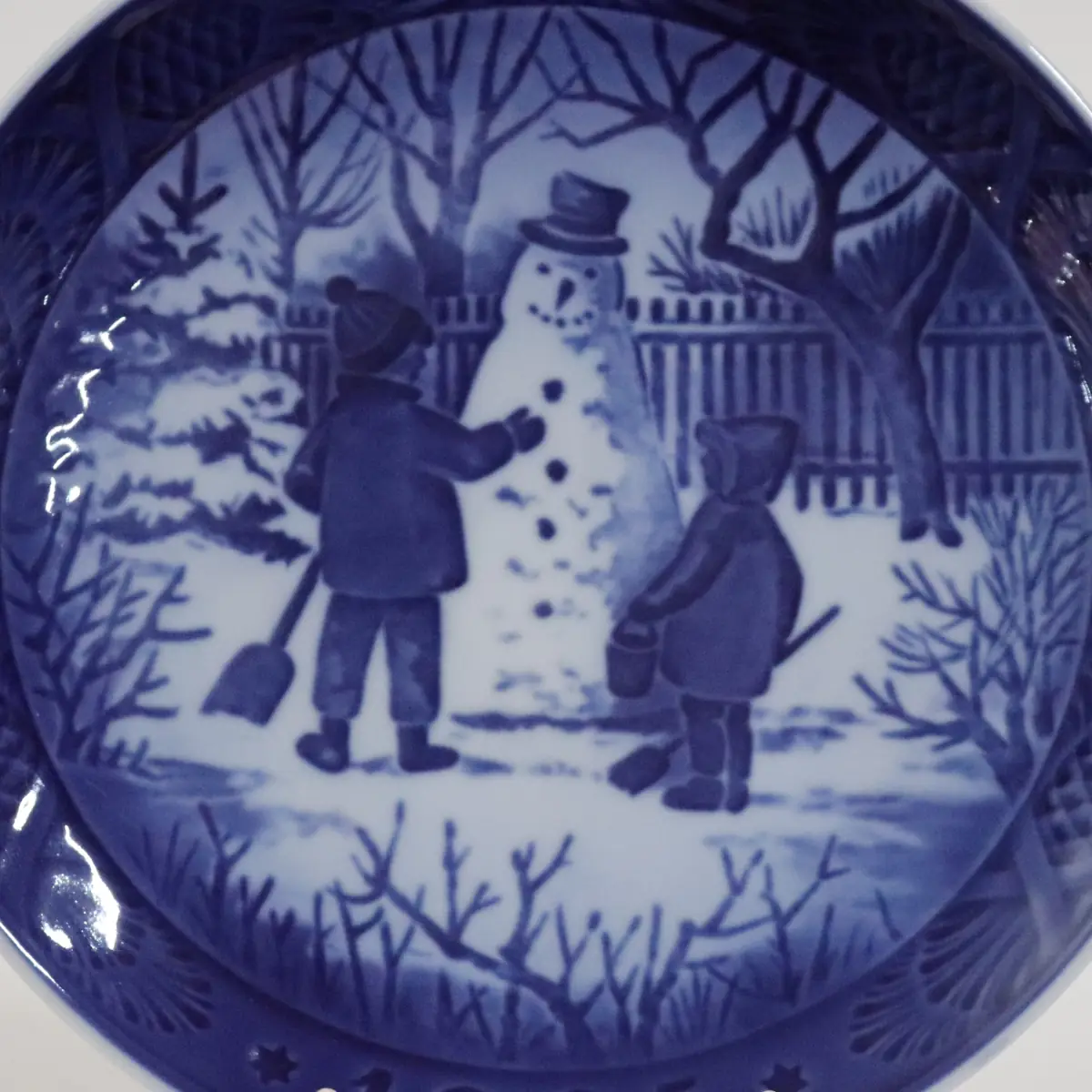 Piatto da collezione in porcellana Royal Copenhagen, Danimarca, decorato in blu sotto smalto. La scena raffigura un idilliaco momento invernale con due bambini intenti a costruire un pupazzo di neve in un giardino innevato. Appartenente alla celebre serie dei piatti natalizi, è apprezzato per il tono ingenuo e poetico, la finezza del disegno e l’elevata qualità esecutiva.