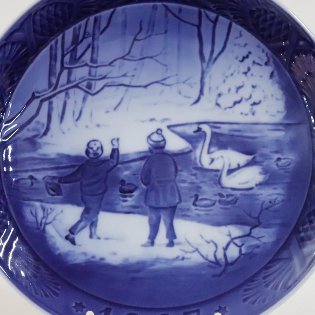Piatto da collezione in porcellana Royal Copenhagen, Danimarca, decorato in blu cobalto sotto smalto. La scena raffigura bambini intenti a nutrire cigni su uno specchio d’acqua invernale, immersi in un paesaggio innevato dal tono poetico. Datato 1987, appartiene alla celebre serie dei piatti natalizi, rinomata per la raffinatezza esecutiva, la qualità della porcellana e il valore collezionistico.