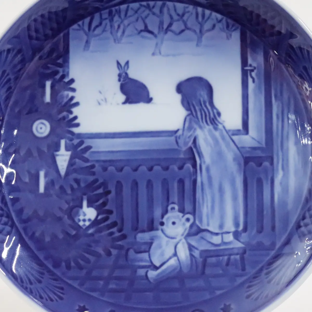 Piatto da collezione in porcellana Royal Copenhagen, Danimarca, decorato in blu sotto smalto. La scena raffigura un interno domestico natalizio: una bambina osserva un coniglio innevato dalla finestra, accanto all’albero di Natale e a un orsacchiotto. Datato 1982, appartiene alla storica serie dei piatti di Natale, apprezzata per il tono intimista, la qualità pittorica e l’ottimo stato di conservazione.