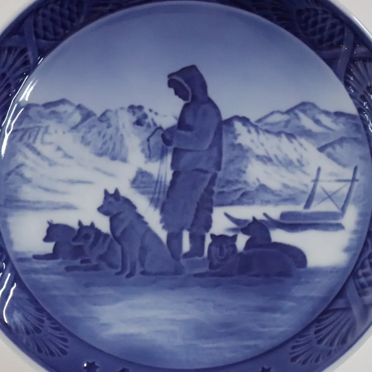 Piatto da collezione in porcellana Royal Copenhagen, Danimarca, decorato in blu sotto smalto. La scena raffigura un paesaggio artico invernale con cacciatore e muta di cani da slitta in sosta, sullo sfondo di montagne innevate. Appartenente alla serie dei piatti natalizi, è apprezzato per l’intensità narrativa e la raffinata resa atmosferica.