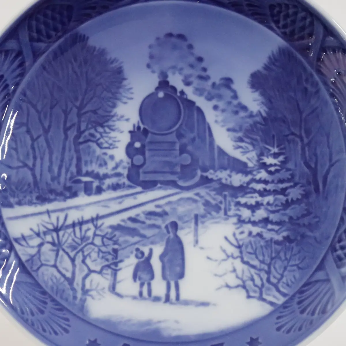 Piatto da collezione in porcellana Royal Copenhagen, Danimarca, decorato in blu sotto smalto. La scena raffigura un paesaggio invernale con locomotiva a vapore in transito e figure di adulti e bambino che osservano il passaggio del treno tra alberi innevati. Appartenente alla tradizione dei piatti natalizi, è apprezzato per il taglio narrativo e la fine resa atmosferica.