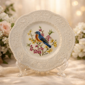 Piatto decorativo in ceramica Bristol Ironstone prodotto dalla manifattura inglese Royal Cauldon, modello “Eastern Bluebird”, con decorazione su disegno di Henry A. Pausch. Il piatto presenta un’elegante tesa ampia con ricca lavorazione a rilievo di gusto classico, a motivi floreali e volute, su fondo avorio leggermente perlato. Al centro è raffigurato un bluebird orientale posato su un ramo fiorito, reso con vivace cromia e buona definizione pittorica, circondato da fiori nei toni del rosa, giallo e verde.