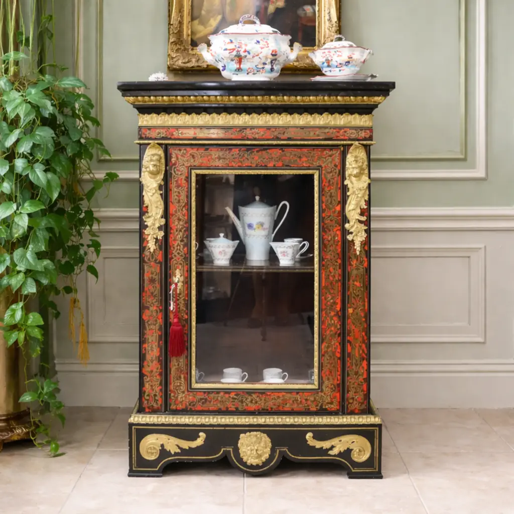 Vetrina alla Boulle in ebano, tartaruga e bronzi dorati Francia, seconda metà del XIX secolo – Stile Napoleone III Raffinata vetrina a un’anta realizzata secondo la celebre tecnica “alla Boulle”, con impiallacciatura in ebano e ricchi intarsi in ottone su fondo in tartaruga rossa naturale, disposti in eleganti motivi a racemi e volute di gusto Luigi XIV. Il prospetto è impreziosito da importanti applicazioni in bronzo dorato finemente cesellato, raffiguranti figure allegoriche laterali, fregi a foglie d’acanto e mascherone centrale nella parte inferiore. L’anta vetrata, incorniciata da profili dorati, apre su ripiani interni destinati all’esposizione di porcellane e oggetti d’arte. Esempio di alta ebanisteria francese del periodo Napoleone III, il mobile coniuga ricchezza ornamentale, equilibrio architettonico e forte impatto scenografico, risultando ideale per ambienti di rappresentanza o collezioni antiquarie di pregio.