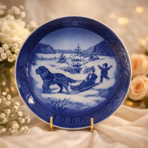 Piatto da parete in porcellana Royal Copenhagen, serie natalizia “Jul på Slottet / Christmas at the Manor House”, anno 1986. Decorazione blu cobalto sottosmalto raffigurante scena invernale con bambini su slitta trainata da cane, entro ampia tesa con motivi stilizzati e data in rilievo. Marchio verde al verso con corona e scritte Royal Copenhagen Denmark. Manifattura danese del XX secolo, in ottimo stato, pezzo da collezione.