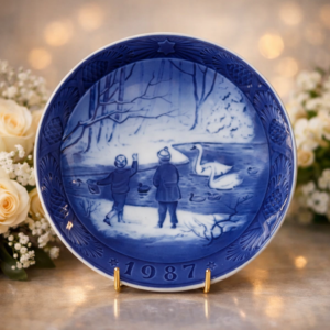 Piatto commemorativo natalizio in porcellana – Royal Copenhagen, 1987 Piatto da collezione in porcellana smaltata a decorazione monocroma blu sottocobalto, appartenente alla serie natalizia annuale della manifattura Royal Copenhagen. Datato 1987 in rilievo sul fronte. La decorazione centrale raffigura una scena invernale all’aperto con due bambini presso uno specchio d’acqua popolato da cigni, ambientata in un paesaggio boschivo innevato, resa con delicate gradazioni tonali di blu e forte senso atmosferico. Bordo decorato a rilievo con motivi vegetali stilizzati e stella simbolica. Al verso, marchio originale Royal Copenhagen con corona reale, tre onde e dicitura Copenhagen – Denmark, con titolo bilingue “Jul på Slottet / Christmas at the Manor House”.