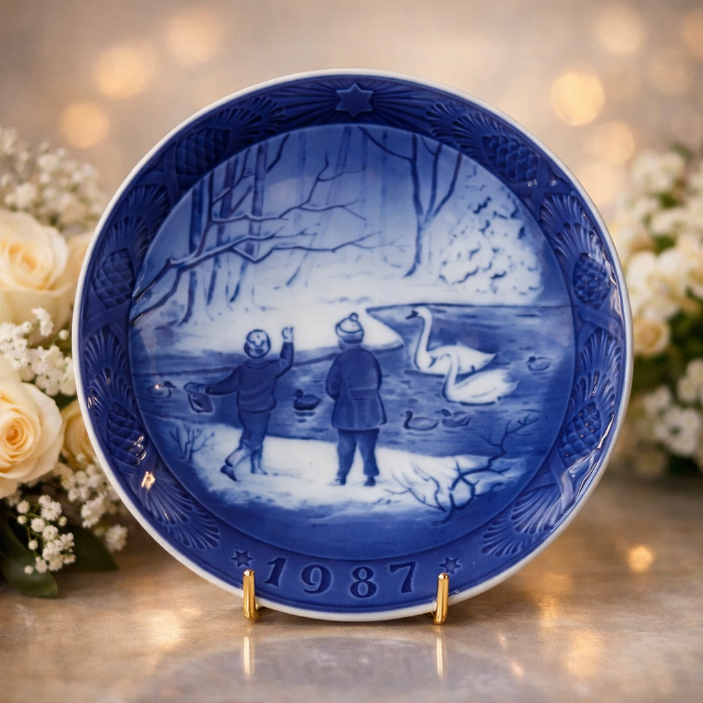 Piatto commemorativo natalizio in porcellana – Royal Copenhagen, 1987 Piatto da collezione in porcellana smaltata a decorazione monocroma blu sottocobalto, appartenente alla serie natalizia annuale della manifattura Royal Copenhagen. Datato 1987 in rilievo sul fronte. La decorazione centrale raffigura una scena invernale all’aperto con due bambini presso uno specchio d’acqua popolato da cigni, ambientata in un paesaggio boschivo innevato, resa con delicate gradazioni tonali di blu e forte senso atmosferico. Bordo decorato a rilievo con motivi vegetali stilizzati e stella simbolica. Al verso, marchio originale Royal Copenhagen con corona reale, tre onde e dicitura Copenhagen – Denmark, con titolo bilingue “Jul på Slottet / Christmas at the Manor House”.