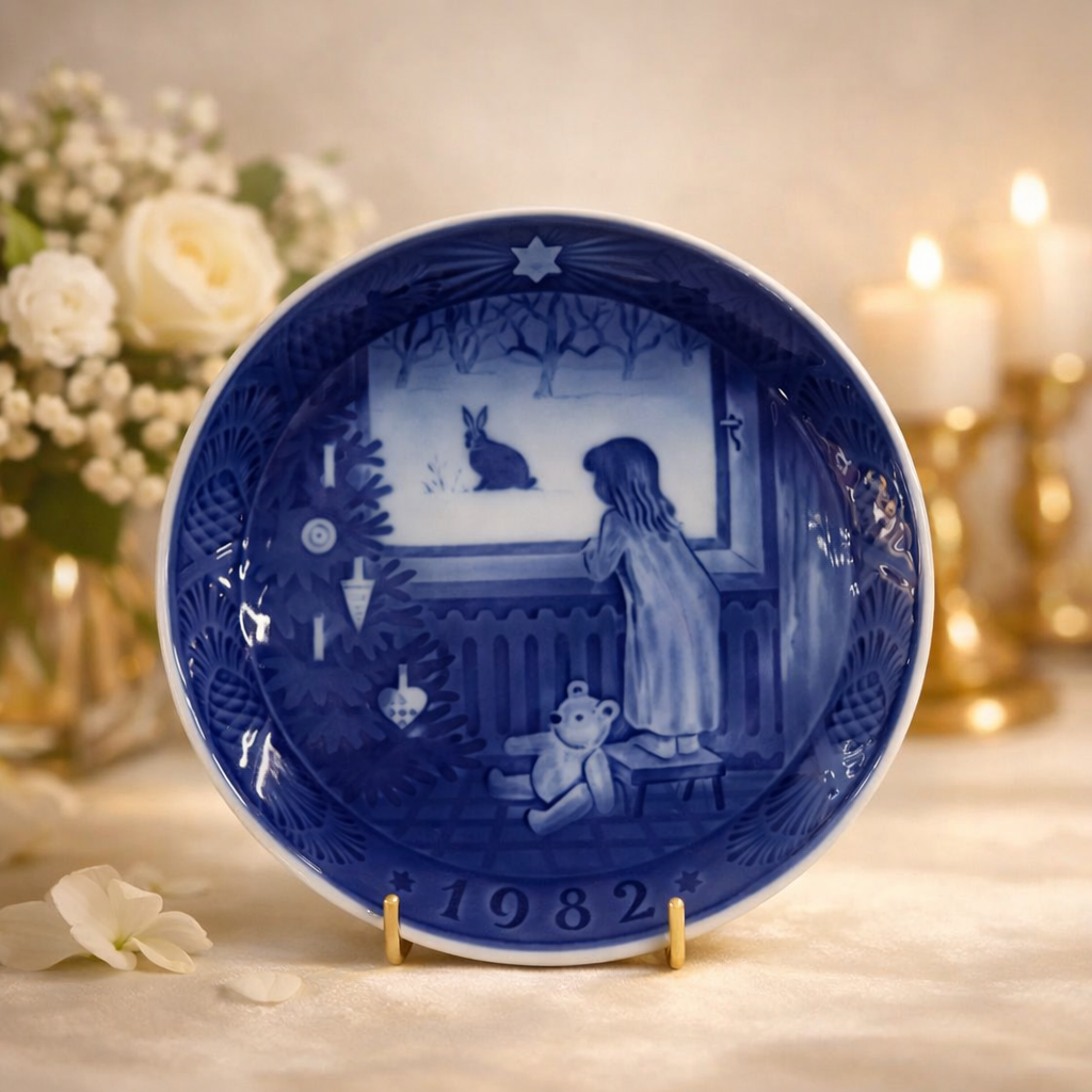 Piatto commemorativo natalizio in porcellana – Royal Copenhagen, 1982 Piatto da collezione in porcellana smaltata a decorazione monocroma blu sottocobalto, appartenente alla serie natalizia annuale della manifattura Royal Copenhagen. Datato 1982 in rilievo sul fronte. La decorazione centrale raffigura una scena d’interno invernale con bambina affacciata alla finestra che osserva un coniglio nella neve, con orsacchiotto in primo piano, resa con delicate gradazioni di blu. Bordo decorato a rilievo con motivi vegetali e stella simbolica. Al verso, marchio originale Royal Copenhagen con corona reale, tre onde e dicitura Copenhagen – Denmark, con titolo bilingue “Jul på Slottet / Christmas at the Manor House”. Stato di conservazione: ottimo.