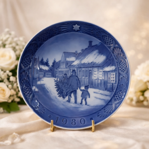 Piatto commemorativo natalizio in porcellana – Royal Copenhagen, 1980 Piatto da collezione in porcellana smaltata a decorazione monocroma blu sottocobalto, appartenente alla serie natalizia annuale della manifattura Royal Copenhagen. Datato 1980 in rilievo sul fronte. La decorazione centrale raffigura una scena invernale rurale con adulto e bambino che trasportano un albero di Natale tra case tradizionali nordiche, resa con eleganti gradazioni di blu. Bordo decorato a rilievo con motivi vegetali e stella simbolica. Al verso, marchio originale Royal Copenhagen con corona reale, tre onde e dicitura Copenhagen – Denmark, con titolo bilingue “Jul på Slottet / Christmas at the Manor House”.