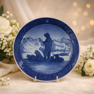Piatto commemorativo natalizio in porcellana – Royal Copenhagen, 1978 Piatto da collezione in porcellana smaltata a decorazione monocroma blu sottocobalto, appartenente alla serie natalizia annuale della manifattura Royal Copenhagen. Datato 1978 in rilievo sul fronte. Decorazione centrale raffigurante una scena artica invernale con conduttore e cani da slitta, ambientata in un paesaggio montano innevato, resa con eleganti gradazioni di blu. Bordo decorato a rilievo con motivi vegetali e stella simbolica. Al verso, marchio originale Royal Copenhagen con corona reale, tre onde e dicitura Copenhagen – Denmark, con titolo bilingue “Jul på Slottet / Christmas at the Manor House”.