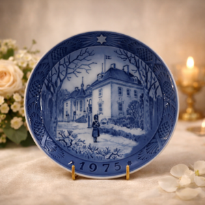 Piatto commemorativo natalizio in porcellana – Royal Copenhagen, 1975 Piatto da collezione in porcellana smaltata a decorazione monocroma blu sottocobalto, realizzato dalla manifattura Royal Copenhagen e appartenente alla serie natalizia annuale. Reca la data 1975 in rilievo sul fronte. La decorazione centrale raffigura una dimora nobiliare immersa in un paesaggio invernale, con sentinella in primo piano, scena di impronta solenne ed elegante resa con raffinate gradazioni di blu. Bordo decorato a rilievo con motivi vegetali e stella simbolica. Al verso, marchio originale Royal Copenhagen con corona reale, tre onde e dicitura Copenhagen – Denmark, con titolo bilingue “Jul på Slottet / Christmas at the Manor House”.