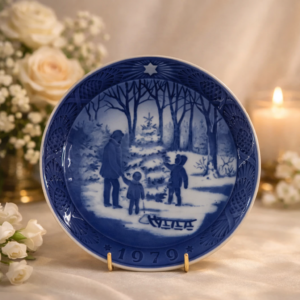 Piatto commemorativo natalizio in porcellana – Royal Copenhagen, 1979 Piatto da collezione in porcellana smaltata a decorazione monocroma blu sottocobalto, prodotto dalla manifattura Royal Copenhagen e appartenente alla serie natalizia annuale. Reca la data 1979 in rilievo sul fronte. La decorazione centrale raffigura un paesaggio boschivo invernale con adulto e due bambini intenti alla scelta dell’albero di Natale, arricchito dalla presenza di una slitta, il tutto reso con eleganti sfumature di blu. Bordo decorato a rilievo con motivi vegetali e stella simbolica. Al verso, marchio originale Royal Copenhagen con corona reale, tre onde e dicitura Copenhagen – Denmark, con titolo bilingue “Jul på Slottet / Christmas at the Manor House”.