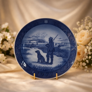 Piatto commemorativo natalizio in porcellana – Royal Copenhagen, 1977 Piatto da collezione in porcellana smaltata a decorazione monocroma blu sottocobalto, realizzato dalla manifattura Royal Copenhagen e appartenente alla serie natalizia annuale. Reca la data 1977 in rilievo sul fronte. La decorazione centrale raffigura un paesaggio rurale invernale con cacciatore e cane, scena di quiete e tradizione resa con raffinate gradazioni di blu. Bordo decorato a rilievo con motivi vegetali e stella simbolica. Al verso, marchio originale Royal Copenhagen con corona reale, tre onde e dicitura Copenhagen – Denmark, con titolo bilingue “Jul på Slottet / Christmas at the Manor House”.
