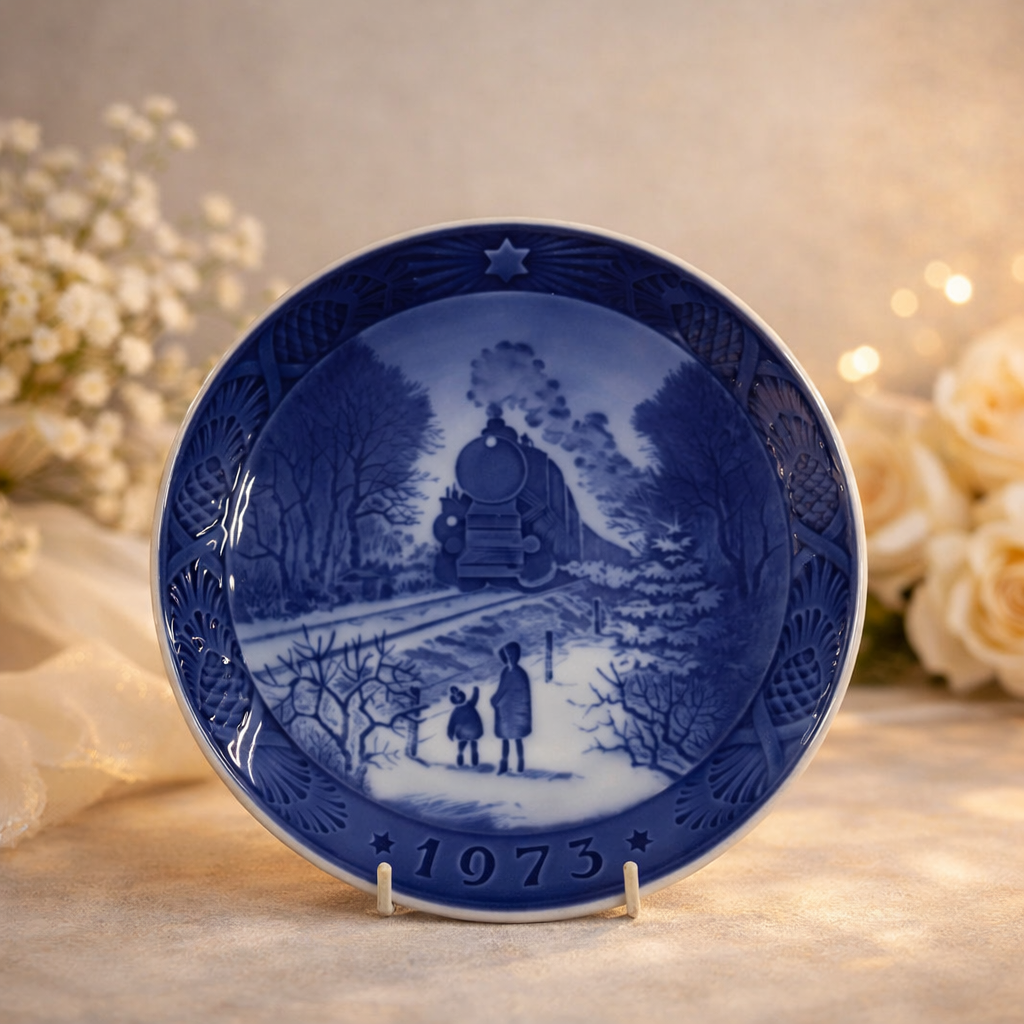 Piatto commemorativo natalizio in porcellana – Royal Copenhagen, 1973 Piatto da collezione in porcellana smaltata a decorazione monocroma blu sottocobalto, prodotto dalla manifattura Royal Copenhagen e appartenente alla serie natalizia annuale. Reca la data 1973 in rilievo sul fronte. La decorazione centrale raffigura un paesaggio invernale con locomotiva a vapore in arrivo e figure in primo piano, scena di forte suggestione narrativa resa con eleganti gradazioni di blu. Bordo decorato a rilievo con motivi vegetali e stella simbolica. Al verso, marchio originale Royal Copenhagen con corona reale, tre onde e dicitura Copenhagen – Denmark, con titolo bilingue “Jul på Slottet / Christmas at the Manor House”.