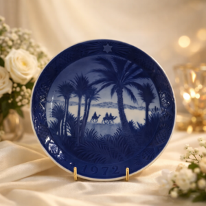 Piatto commemorativo natalizio in porcellana – Royal Copenhagen, 1972 Piatto da collezione in porcellana smaltata con decorazione monocroma blu sottocobalto, realizzato dalla manifattura Royal Copenhagen. Appartenente alla storica serie natalizia annuale, reca la data 1972 in rilievo sul fronte. La scena centrale raffigura un paesaggio orientale con palme e carovana di cammelli nel deserto, interpretazione evocativa del tema natalizio, eseguita con finezza grafica e forte senso atmosferico. Bordo decorato con motivi vegetali stilizzati e stella simbolica. Al verso, marchio originale Royal Copenhagen con corona reale, tre onde e dicitura Copenhagen – Denmark, accompagnato dal titolo bilingue “Jul på Slottet / Christmas at the Manor House”.
