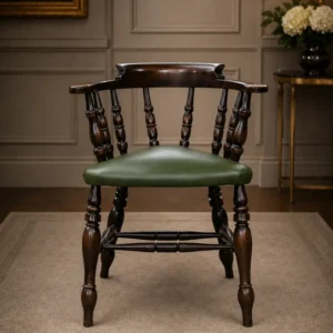 Elegante poltroncina inglese di gusto vittoriano, appartenente alla tipologia Captain Chair, realizzata in legno massello con raffinata struttura a colonnine tornite e schienale avvolgente con braccioli integrati. La seduta imbottita è rivestita in pelle verde con bordura decorativa a borchie, dettaglio tipico dell’arredo britannico da studio e biblioteca. Le gambe tornite unite da traverse sagomate conferiscono solidità e armonia alla struttura, mentre la calda patina del legno ne esalta il fascino autentico. In buono stato conservativo generale, è ideale come sedia da scrittoio, poltroncina da lettura o complemento d’arredo per ambienti classici ed eleganti.