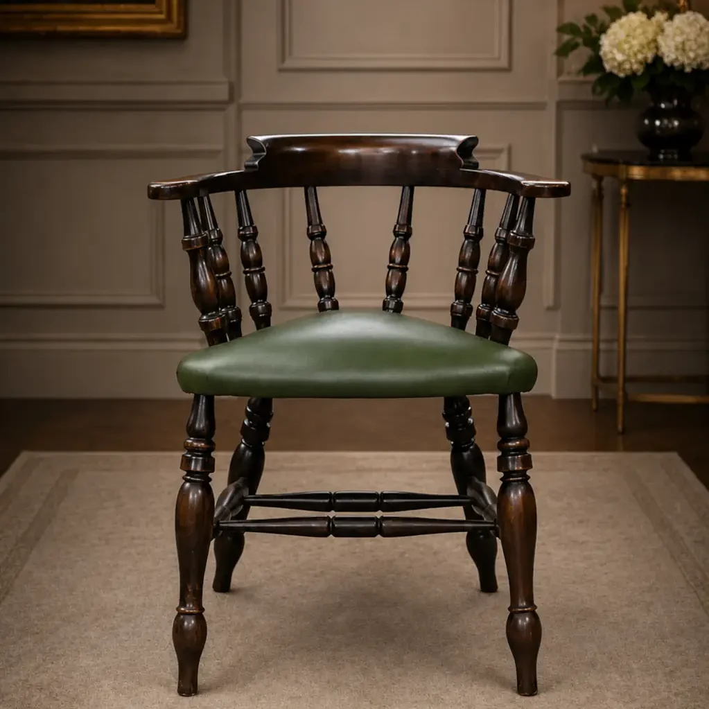 Elegante poltroncina inglese di gusto vittoriano, appartenente alla tipologia Captain Chair, realizzata in legno massello con raffinata struttura a colonnine tornite e schienale avvolgente con braccioli integrati. La seduta imbottita è rivestita in pelle verde con bordura decorativa a borchie, dettaglio tipico dell’arredo britannico da studio e biblioteca. Le gambe tornite unite da traverse sagomate conferiscono solidità e armonia alla struttura, mentre la calda patina del legno ne esalta il fascino autentico. In buono stato conservativo generale, è ideale come sedia da scrittoio, poltroncina da lettura o complemento d’arredo per ambienti classici ed eleganti.