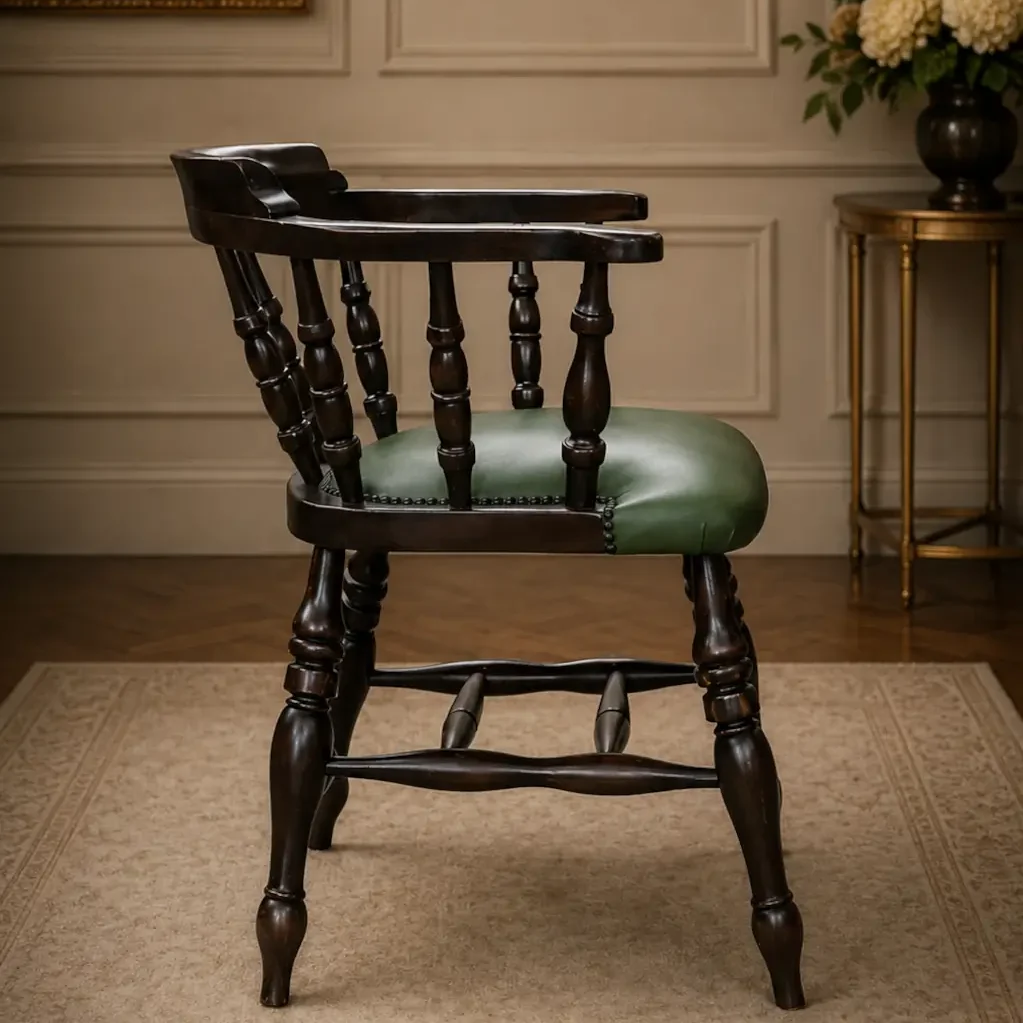Elegante poltroncina inglese di gusto vittoriano, appartenente alla tipologia Captain Chair, realizzata in legno massello con raffinata struttura a colonnine tornite e schienale avvolgente con braccioli integrati. La seduta imbottita è rivestita in pelle verde con bordura decorativa a borchie, dettaglio tipico dell’arredo britannico da studio e biblioteca. Le gambe tornite unite da traverse sagomate conferiscono solidità e armonia alla struttura, mentre la calda patina del legno ne esalta il fascino autentico. In buono stato conservativo generale, è ideale come sedia da scrittoio, poltroncina da lettura o complemento d’arredo per ambienti classici ed eleganti.