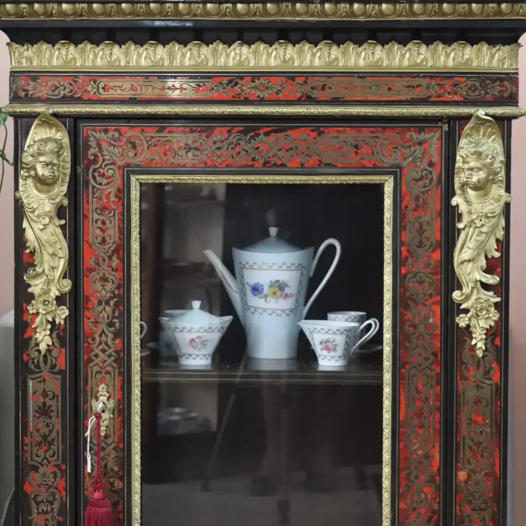 Vetrina alla Boulle in ebano, tartaruga e bronzi dorati Francia, seconda metà del XIX secolo – Stile Napoleone III Raffinata vetrina a un’anta realizzata secondo la celebre tecnica “alla Boulle”, con impiallacciatura in ebano e ricchi intarsi in ottone su fondo in tartaruga rossa naturale, disposti in eleganti motivi a racemi e volute di gusto Luigi XIV. Il prospetto è impreziosito da importanti applicazioni in bronzo dorato finemente cesellato, raffiguranti figure allegoriche laterali, fregi a foglie d’acanto e mascherone centrale nella parte inferiore. L’anta vetrata, incorniciata da profili dorati, apre su ripiani interni destinati all’esposizione di porcellane e oggetti d’arte. Esempio di alta ebanisteria francese del periodo Napoleone III, il mobile coniuga ricchezza ornamentale, equilibrio architettonico e forte impatto scenografico, risultando ideale per ambienti di rappresentanza o collezioni antiquarie di pregio.